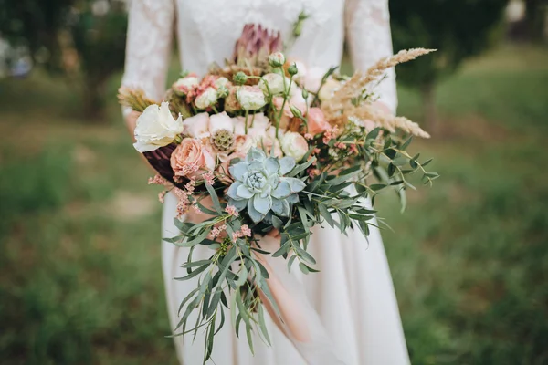 Pengantin dalam gaun berdiri di kebun hijau dan memegang buket pengantin bunga dan hijau Stok Gambar