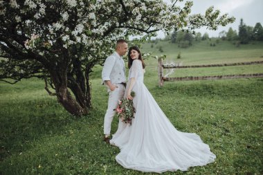 Dağlarda bahar düğünü. Beyaz gömlekli ve pantolonlu genç bir adam ve beyaz elbiseli bir kız elinde bir buket kırmızı çiçekle bir çiçeğin altında duruyorlar.