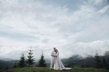 Dağlarda bahar düğünü. Beyaz gömlekli ve pantolonlu genç bir adam ve beyaz elbiseli bir kız gökyüzü ve ağaçların arka planında dağın zirvesinde dikiliyorlar.
