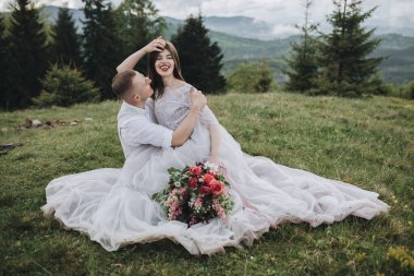 Dağlarda bahar düğünü. Beyaz gömlekli ve pantolonlu genç bir adam ve beyaz elbiseli bir kız dağlardaki yeşil çimlerin üzerinde oturuyorlar ve ellerinde bir buket kırmızı çiçek var.