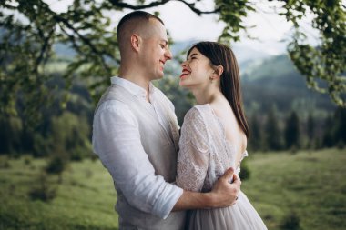 Dağlarda bahar düğünü. Beyaz gömlekli ve pantolonlu genç bir adam ve beyaz elbiseli bir kız gün batımında dağlarda büyük bir ağacın dallarının altında duruyorlar.