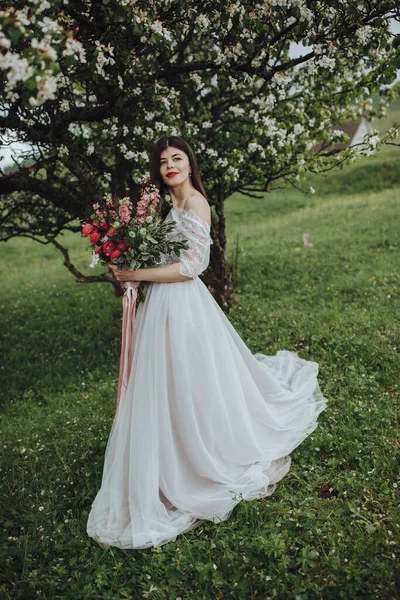 山の中で春の結婚式 白いドレスの少女が山の中の花の木の近くに立って リボンで赤い花の花束を手に持っています — ストック写真