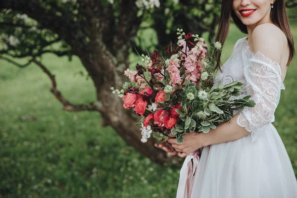 山の中で春の結婚式 白いドレスの少女が山の中の花の木の近くに立って リボンで赤い花の花束を手に持っています — ストック写真