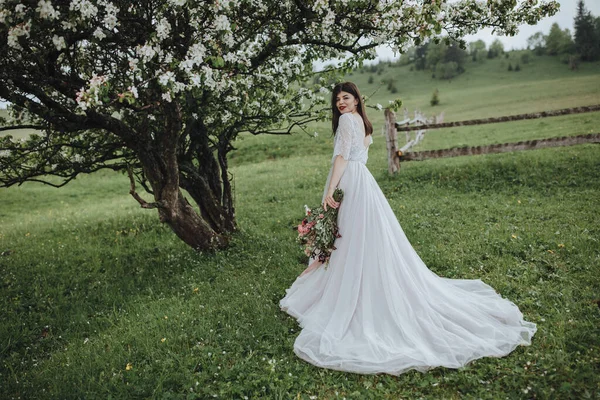 山の中で春の結婚式 白いドレスの少女が山の中の花の木の近くに立って リボンで赤い花の花束を手に持っています — ストック写真