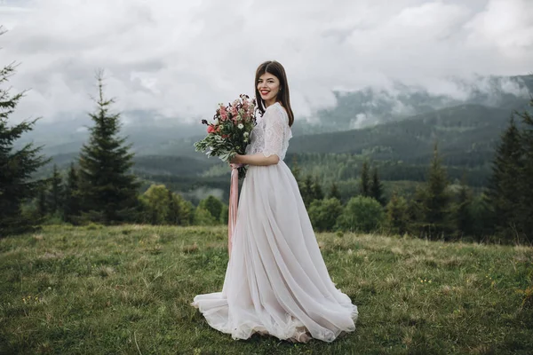 山の中で春の結婚式 山の緑の草原に白い服を着た少女が立っていて 手には赤い花と緑の花束を持っている — ストック写真