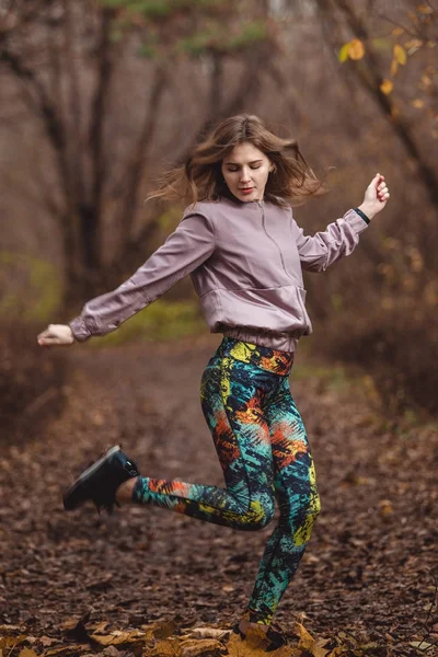 Portrait of sporty girl in white shirt performs warming-up before jogging in autumn forest.