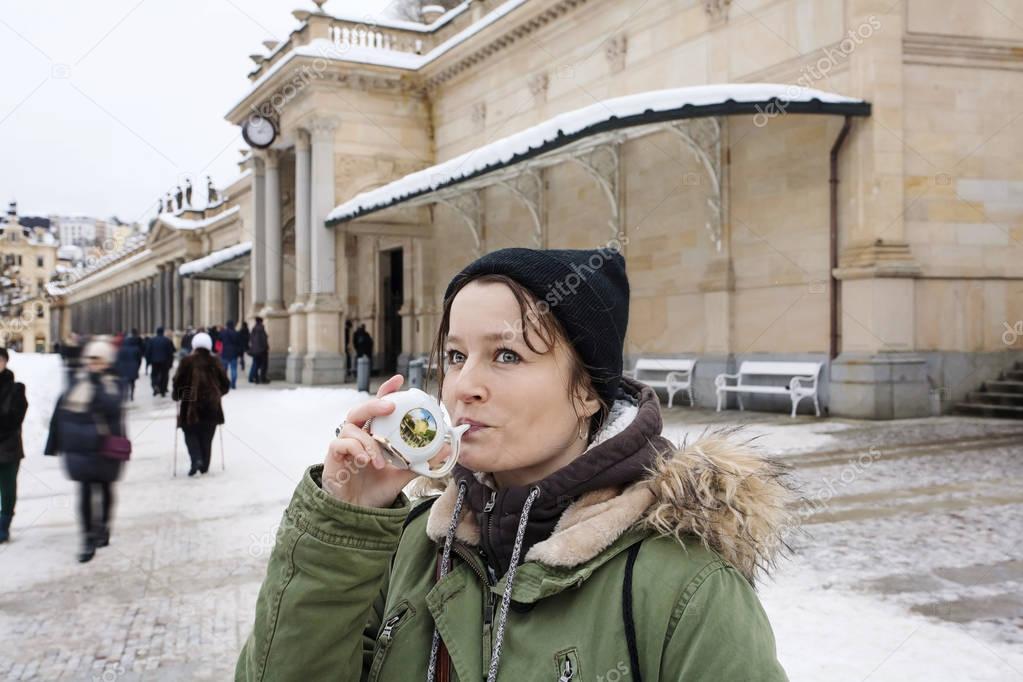Young woman drinking from cup with therapeutic mineral water at a natural hot spring in Karlovy Vary during winter time, Czech Republic