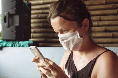 Caucasian female tourist with face mask in India with her cellphone just realized, that her flight was cancelled clipart