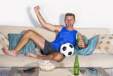 genç çekici adam mutlu ve heyecanlı izlerken futbol maçı televizyonda zafer hedef deli ve spastik bira patlamış mısır ve futbol topu ile kutluyor 