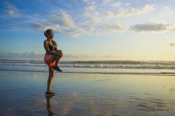 Joven atractiva mujer asiática estiramiento de la pierna en el calentamiento antes de ejecutar entrenamiento en la playa puesta del sol con increíble hermoso reflejo de la luz del sol en la arena — Foto de Stock