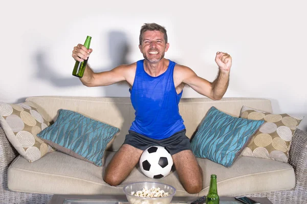 Joven atractivo hombre feliz y emocionado viendo el partido de fútbol en la televisión celebrando la victoria gol loco y espástico con palomitas de cerveza y pelota de fútbol — Foto de Stock