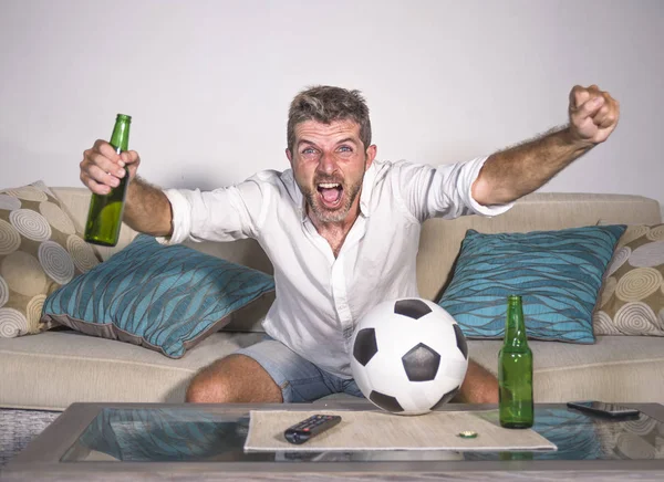Joven atractivo hombre feliz y emocionado viendo el partido de fútbol en la televisión celebrando la victoria gol loco y espástico con palomitas de cerveza y pelota de fútbol — Foto de Stock