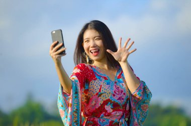 genç güzel ve mutlu Asyalı Çinli turist kadına selfie pic alarak hareket eden telefon fotoğraf makinesi ile tropikal sahada renkli elbise ile 20'li