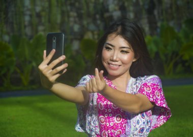 yeşil çim park açık havada el parmak işaretiyle aşk yaparken cep telefonu kamera ile selfie pic alarak genç mutlu ve güzel Asyalı Çinli kadın 