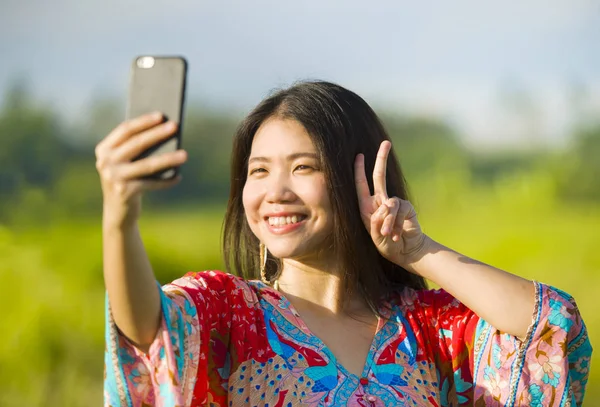 Jovem bonita e feliz asiática chinesa turista mulher em seus 20s com vestido colorido tendo selfie pic com câmera de telefone móvel no campo tropical — Fotografia de Stock