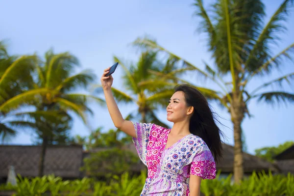 Happy beautiful and gorgeous Asian Chinese woman in glamour dress taking self portrait selfie photo with mobile phone at holidays tropical resort — Stock Photo, Image