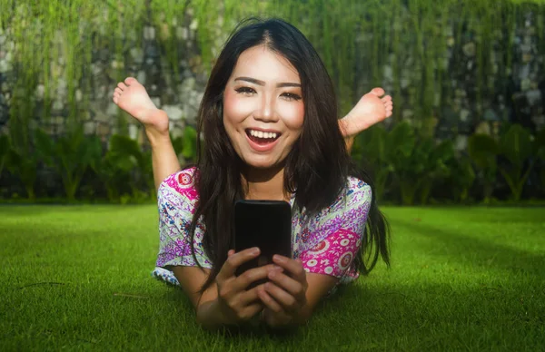 Jonge aantrekkelijke mooie en prachtige Aziatische Chinese vrouw liggend op groen gras park met behulp van internet sociale media app op mobiele telefoon ontspannen en gelukkig — Stockfoto