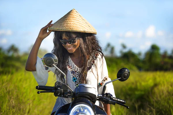 Young beautiful tourist or nomad traveler black afro American woman riding motorbike in tropical field wearing traditional Asian hat smiling happy — Stock Photo, Image