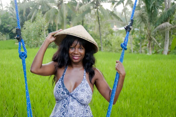 Young beautiful tourist afro american black woman riding swing enjoying and smiling happy wearing traditional Asian hat in jungle and rice field tropical — Stock Photo, Image