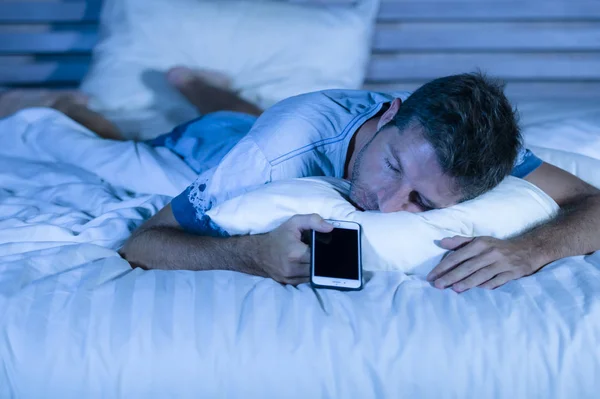 Aantrekkelijke moe man in bed in slaap vallen tijdens het gebruik van de mobiele telefoon nog steeds houdt de cellulaire in zijn hand tijdens het slapen in internet en sociale media-verslaving — Stockfoto