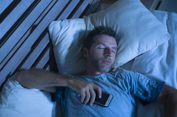 Attractive tired man in bed falling asleep while using mobile phone still holding the cellular in his hand while sleeping in internet and social media addiction — Stock Photo, Image