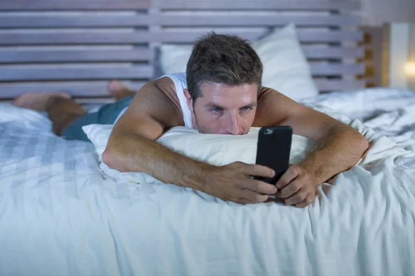 Портрет молодого привлекательного мужчины, лежащего в постели уставшего и расслабленного с помощью мобильного телефона, выглядящего сонным в своей спальне в интернет-зависимости — стоковое фото