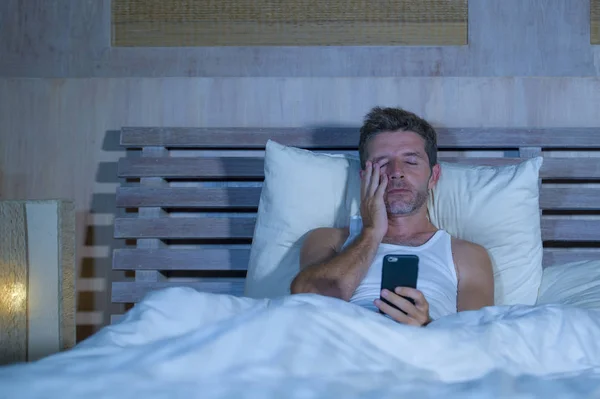 Retrato de un hombre joven y atractivo tumbado en la cama cansado y relajado usando el teléfono móvil mirando somnoliento en su dormitorio de apartamento en la adicción a Internet —  Fotos de Stock