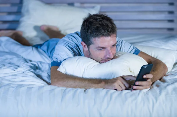Joven atractivo y feliz hombre acostado en la cama usando internet teléfono móvil sonriendo enviar texto en las redes sociales y la adicción a la comunicación celular —  Fotos de Stock