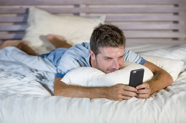 Молодой привлекательный и счастливый мужчина лежит на кровати с помощью интернет-телефона улыбаясь отправка текста в социальных сетях и сотовой связи наркомании — стоковое фото