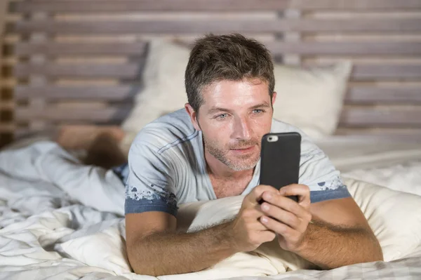 Joven atractivo y feliz hombre acostado en la cama usando internet teléfono móvil sonriendo enviar texto en las redes sociales y la adicción a la comunicación celular — Foto de Stock