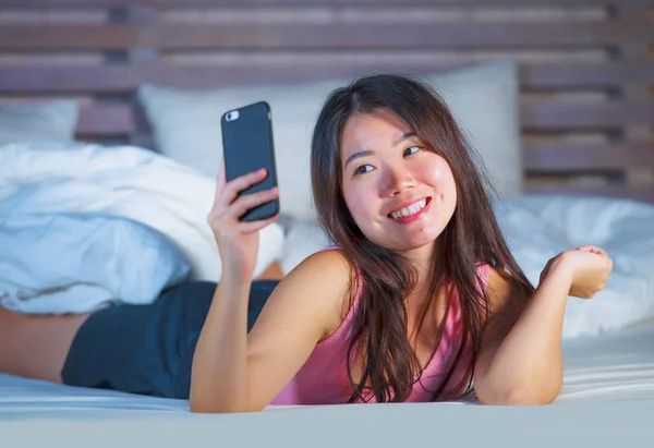 Молодая красивая и счастливая азиатская китаянка в возрасте 20 лет фотографирует себя с мобильного телефона, улыбаясь или делая видео-звонок на свидании лежа на кровати — стоковое фото