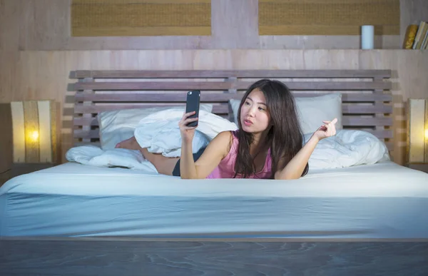 Молодая красивая и счастливая азиатская китаянка в возрасте 20 лет фотографирует себя с мобильного телефона или делает видео-звонок, лежа на кровати ночью — стоковое фото