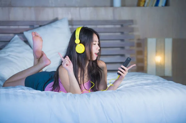 Joven atractivo y feliz asiático chino mujer con auriculares amarillos escuchar música en el teléfono móvil en la cama en casa sonriendo divertirse — Foto de Stock