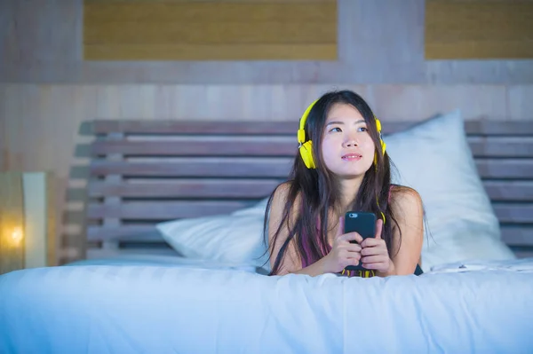 Jovem atraente e feliz mulher chinesa asiática com fones de ouvido amarelos ouvir música no celular na cama em casa sorrindo se divertindo — Fotografia de Stock