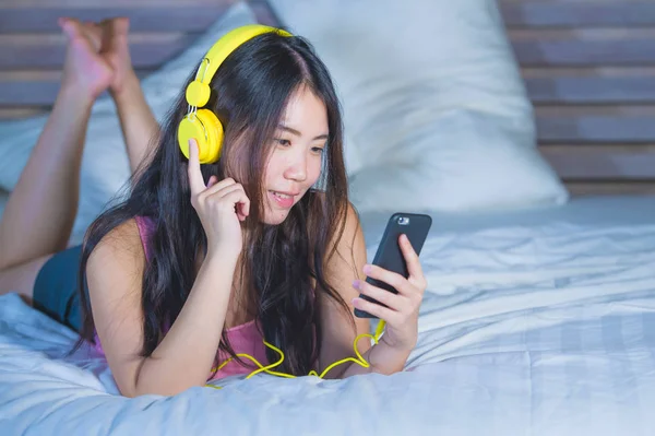Genç çekici ve mutlu Asya Çin sarı kulaklıklarla müzik cep telefonu evde yatakta eğlence gülümseyen kadın — Stok fotoğraf