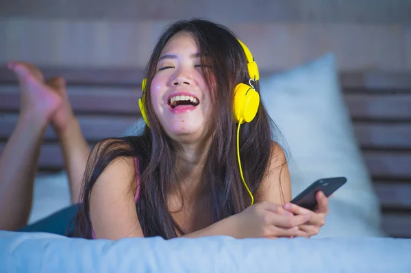 Genç çekici ve mutlu Asya Çin sarı kulaklıklarla müzik cep telefonu evde yatakta eğlence gülümseyen kadın — Stok fotoğraf