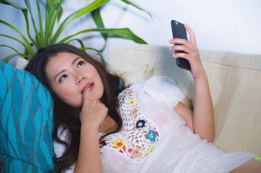 Internet cep telefonuyla muhteşem görünümlü genç güzel ve 20'li veya oturma odası kanepe yalan 30s mutlu Asyalı Çinli kadın kanepe