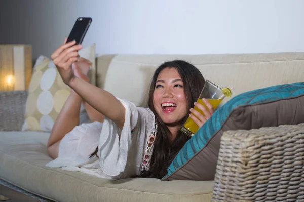 Jeune belle et heureuse asiatique chinois 20s ou 30s femme prendre selfie photo avec téléphone mobile boire du jus d'orange à la maison canapé salon — Photo