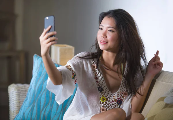 young beautiful and happy Asian Chinese 20s or 30s woman taking selfie picture with mobile phone sitting at home living room sofa couch smiling