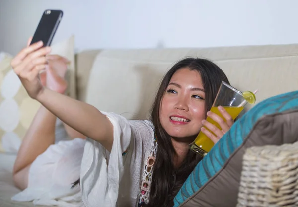 Jeune belle et heureuse asiatique chinois 20s ou 30s femme prendre selfie photo avec téléphone mobile boire du jus d'orange à la maison canapé salon — Photo