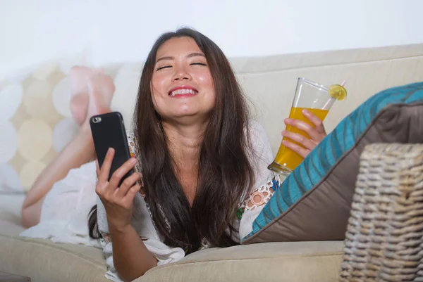 Jeune belle et heureuse asiatique chinois femme sur son 20s ou 30s couché au canapé salon de boire du jus d'orange sain en utilisant Internet mobile — Photo