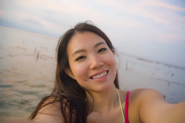 Молодая счастливая и красивая азиатская туристка-кореянка делает автопортретное селфи с мобильным телефоном, улыбаясь и веселясь на пляже заката — стоковое фото