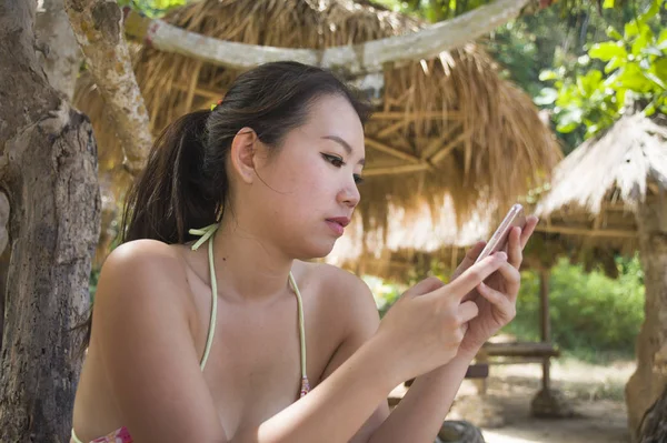 Jovem bonita e feliz mulher coreana asiática em biquíni usando a internet no telefone celular no paraíso tropical praia resort sob sombra palmeiras — Fotografia de Stock