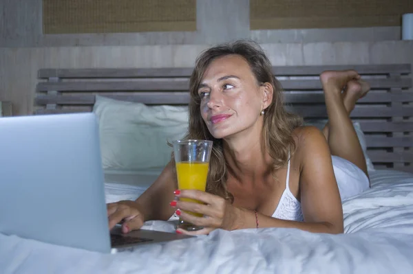 Красивая белая женщина 30 лет лежит на кровати ночью в спальне дома с помощью интернета на ноутбуке пить здоровый апельсиновый сок — стоковое фото