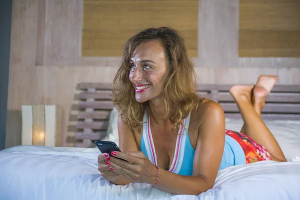 Молодая привлекательная и красивая женщина дома в постели с помощью интернет приложения социальных медиа на мобильном телефоне улыбаясь счастливый пить здоровый апельсиновый сок расслабленной — стоковое фото