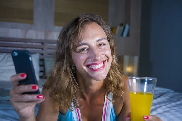 Молодая привлекательная и красивая женщина дома в постели с помощью интернет приложения социальных медиа на мобильном телефоне улыбаясь счастливый пить здоровый апельсиновый сок расслабленной — стоковое фото