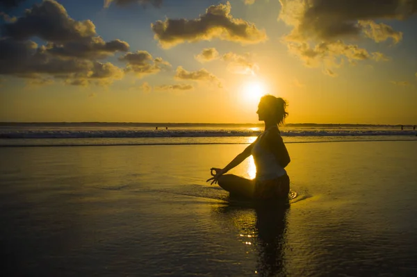 silhouette of young fit and attractive sport woman in beach sunset yoga practice workout sitting on wet sun in front of sea in meditation relaxation