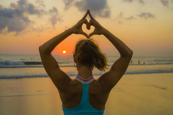 Retrato de espalda de mujer joven en forma y atractiva saludable haciendo amor en forma de corazón con manos y dedos contra la increíble hermosa playa de atardecer en yoga — Foto de Stock