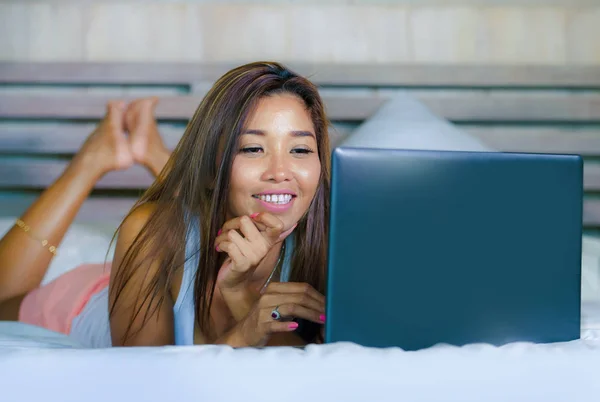 Молодая красивая и счастливая азиатская латиноамериканка смешанной расы улыбается в спальне дома, лежа на кровати с помощью Интернета на ноутбуке расслабленной и прохладной — стоковое фото