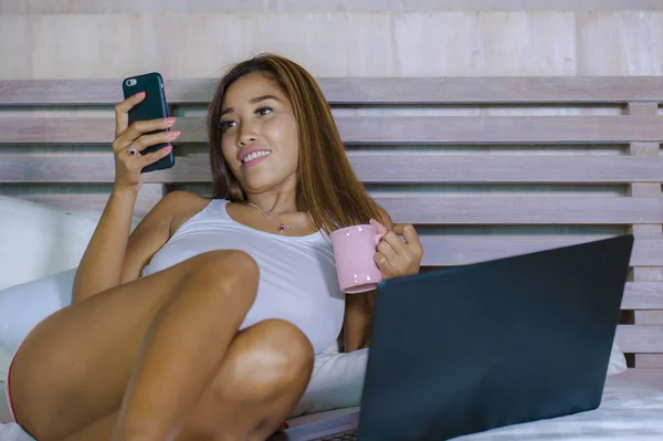 Jonge mooie en gelukkig Aziatische Hispanic gemengd ras vrouw die lacht op huis slaapkamer liggend op bed met behulp van computer laptop verzenden van tekst op mobiele telefoon — Stockfoto